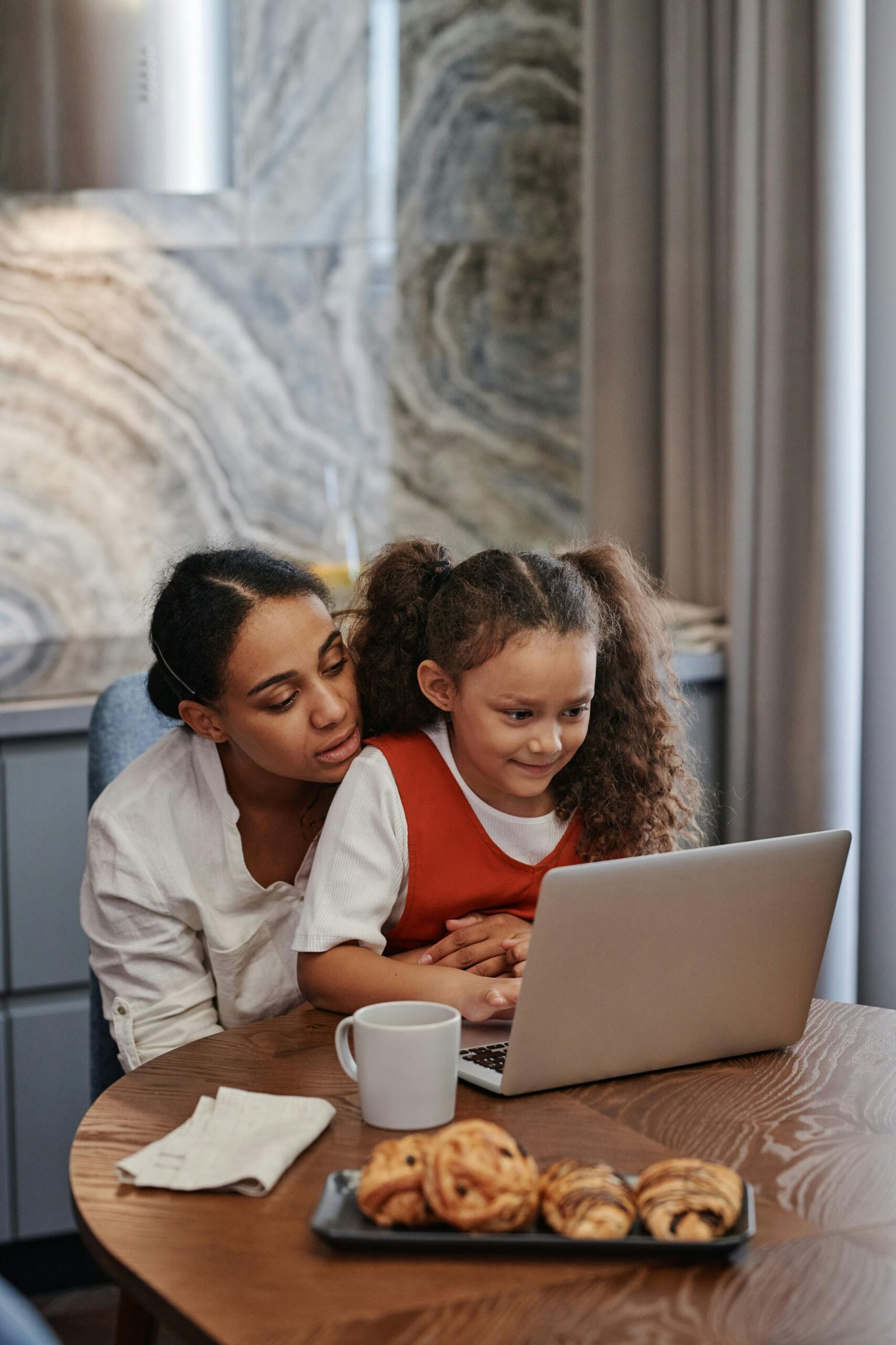 A mother and daughter smiling while using a laptop for online learning at home.